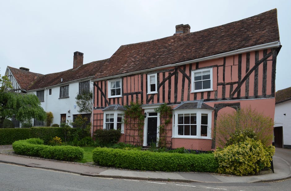 A row of three houses on a residential street, with the central house featuring a distinctive pink exterior with black timber framing, characteristic of period homes. The pink house has a white front door and white-framed sash windows, with some greenery, including climbing plants, growing along the facade. In front of the houses, there is a well-maintained garden with low, neatly trimmed hedges, small bushes, and a flowering shrub on the right. A paved pathway runs parallel to the curb, leading to the front door of the pink house. The street is paved with asphalt, and the sky overhead is overcast, suggesting typical house removal or relocation activities, with the potential for furniture transport or packing operations nearby. The scene reflects a residential setting suitable for professional house removals aimed at avoiding damage to period homes in Ponders End, as managed by Man with Van Ponders End.