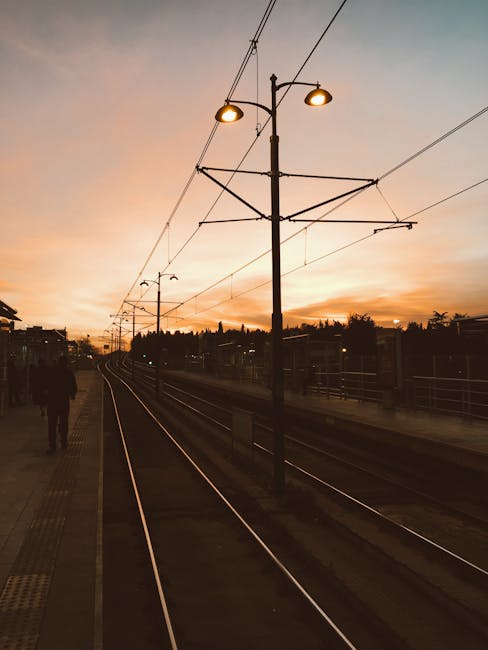 A view of the train tracks at Ponders End station during sunset, with overhead electric wires and supporting poles lining the platform. The sky is tinged with warm hues of orange and yellow, casting a soft glow over the scene. On the left side of the image, a person is seen walking along the platform, which is equipped with tactile paving for safety. In the background, there are silhouetted trees and buildings, suggesting an urban residential area nearby. The scene captures the outdoor environment of a typical UK railway station, emphasizing the infrastructure involved in house removals and property relocation services, as provided by Man with Van Ponders End. Such infrastructure supports efficient furniture transport and home relocation logistics, especially around the Ponders End and Meridian Water area.