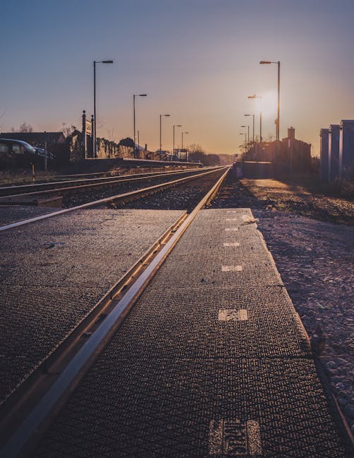 A street-level view of a railway crossing during sunset, with multiple sets of train tracks running parallel into the distance. The crossing features a textured rubber or composite surface with white safety markings along the edge, and it is bordered by patches of gravel and pavement. Tall streetlights line both sides of the tracks, some with illuminated fixtures, and a few vehicles are parked or visible in the background near residential houses with sloped roofs on the left side of the image. The sky is clear with a warm glow from the setting sun, casting long shadows across the scene. This setting depicts a typical location where home relocation or furniture transport services by Man with Van Ponders End could be coordinated, especially during the loading process of a house move involving careful navigation through local routes from Ponders End station to nearby areas such as Meridian Water.