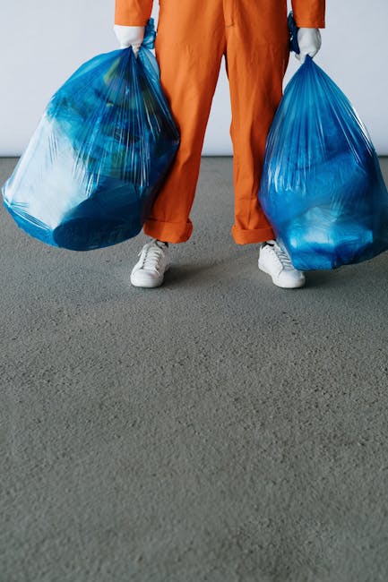 A person wearing orange overalls and white sneakers is standing on a grey carpeted floor, holding two large blue plastic bags filled with bulky waste items for house clearance. The individual’s hands, covered in grey gloves, grip the bags by their handles, with one bag in each hand. The background shows a plain white wall, and part of a doorway is visible behind the person, suggesting an indoor setting suitable for packing or moving operations. This scene illustrates the process of load preparation and waste removal within a property, relevant to home relocation and furniture transport services provided by Man with Van Ponders End, especially in relation to bulky waste removals or packing and moving activities.