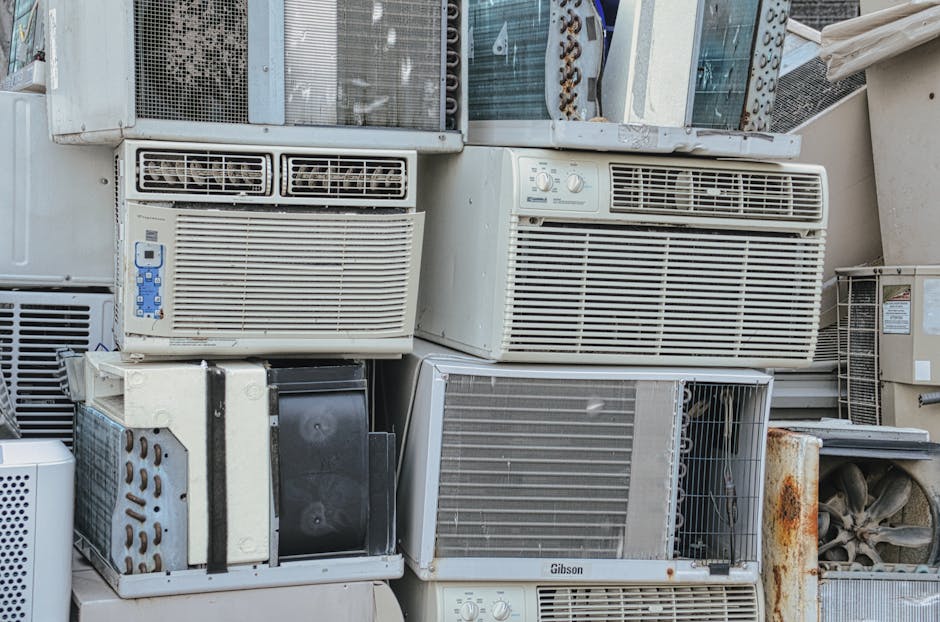 A large pile of various used and discarded air conditioning units and dehumidifiers stacked outdoors on a concrete surface; some units display rust, dirt, and visible wear. The units are predominantly white or beige, with different sizes and styles, including window and portable types, with vents, control panels, and grilles visible. They are arranged in a haphazard manner, with some on top of others, creating a cluttered stack. The setting appears to be behind a building or in a storage area, with natural daylight illuminating the scene. This image showcases the disposal or clearing of bulky waste, illustrating the kind of items transported during home relocation or bulk waste removals carried out by Man with Van Ponders End, who specialise in furniture transport and packing services.