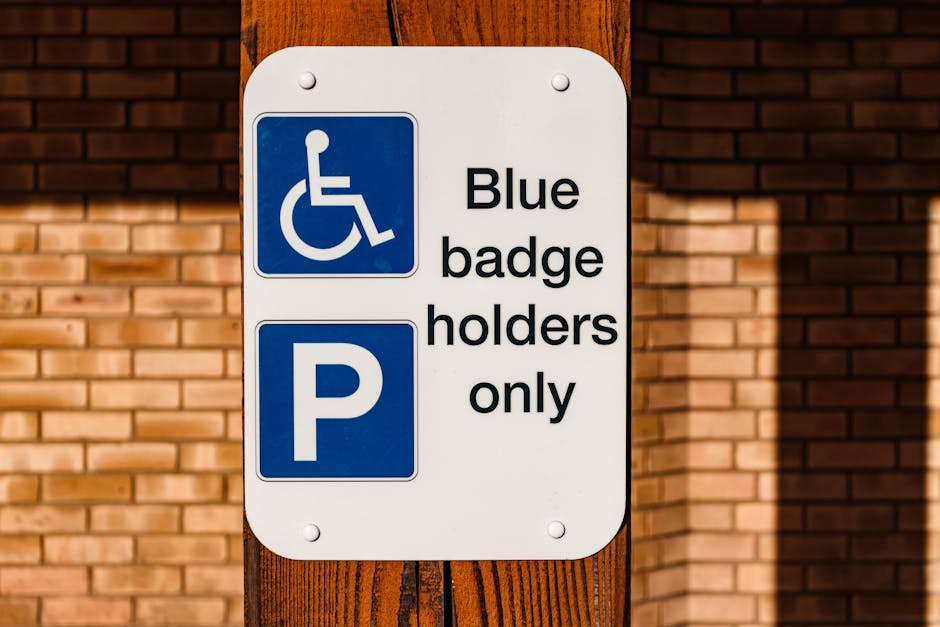 A close-up photograph of a rectangular white sign mounted on a wooden post situated on a pavement in front of a brick wall. The sign features a blue accessibility symbol for wheelchair users at the top, indicating designated parking for disabled badge holders. Below the symbol, there is black text reading 'Blue badge holders only,' and underneath, a large blue P symbol indicating parking availability. The setting suggests an outdoor area near a property entrance or loading zone, relevant to house removals or loading processes. The sign’s purpose is to inform drivers that parking is reserved exclusively for those with a valid blue badge, which is important for planning home relocation and furniture transport logistics. The lighting is natural, highlighting the sign’s clarity and the texture of the brick wall behind it.