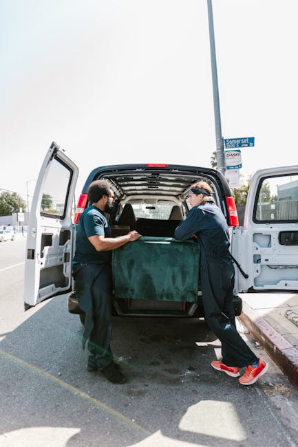 Two movers from Man with Van Ponders End are loading a large, dark green upholstered piece of furniture into the back of a black van during a home relocation process on a pavement in Ponders End. The van's rear doors are wide open, revealing the interior with a few cardboard boxes and packing materials. One mover, wearing a navy jacket and red sneakers, is holding the furniture while the other, dressed in a dark shirt, assists with positioning it inside the vehicle. The scene takes place outdoors under bright daylight, with a street sign visible on a nearby lamppost and a clear sky overhead. This loading activity is part of a furniture transport and packing process involved in a residential move, supported by the professional team from Man with Van Ponders End, ensuring careful handling of belongings for effective home relocation and moving logistics.