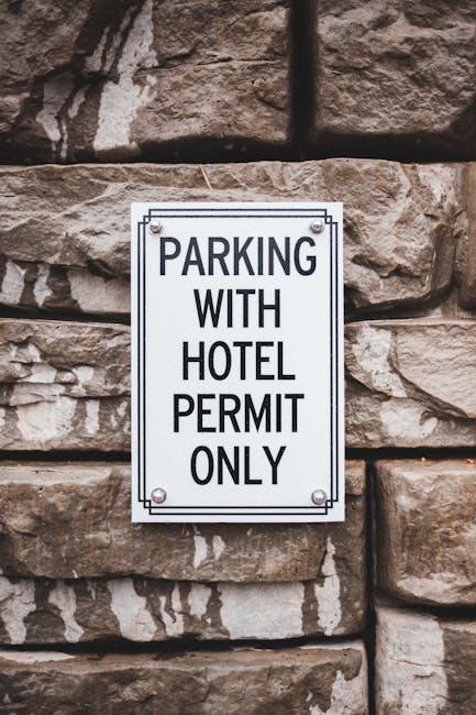 A close-up photograph of a rectangular white sign mounted on a wooden post situated on a pavement in front of a brick wall. The sign features a blue accessibility symbol for wheelchair users at the top, indicating designated parking for disabled badge holders. Below the symbol, there is black text reading 'Blue badge holders only,' and underneath, a large blue P symbol indicating parking availability. The setting suggests an outdoor area near a property entrance or loading zone, relevant to house removals or loading processes. The sign’s purpose is to inform drivers that parking is reserved exclusively for those with a valid blue badge, which is important for planning home relocation and furniture transport logistics. The lighting is natural, highlighting the sign’s clarity and the texture of the brick wall behind it.