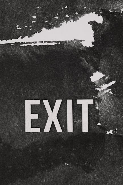 Close-up view of a black, distressed exterior wall with a partially worn white paint marking, displaying the word 'EXIT' in bold, uppercase letters. The surface shows peeling paint and patches of exposed material, indicating weathered outdoor conditions. In the background, a section of pavement or ground surface is visible, suggesting the location is near a building's exit point. This image relates to home or building evacuation signage, possibly during a property move or emergency situation, aligning with the services provided by Man with Van Ponders End for removals and logistical support.