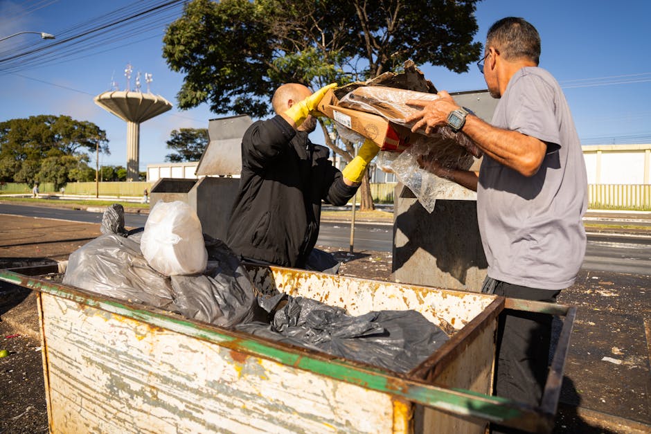 Two men are engaged in a home relocation process involving waste disposal. They are standing outside near a large, open metal skip filled with black and white rubbish bags and various pieces of packaging material, including plastic wrap and cardboard boxes. One man, wearing a black jacket and yellow gloves, is placing a cardboard box into the skip, while the other, dressed in a light grey T-shirt, holds and supports the box. The scene takes place on a paved outdoor area with clear weather, and in the background, there are trees, utility poles, and a white building. The image captures the loading process during a move, with tools such as straps or trolleys not visible, but the focus is on the careful handling of waste, reflecting proper disposal practices in a move handled by Man with Van Ponders End, aligned with their removals and disposal services as outlined in the Enfield Council Waste Rules page.