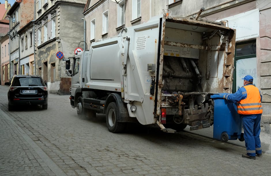 A sanitation worker dressed in a high-visibility orange vest and blue uniform is shown standing on a cobbled residential street, emptying a large blue wheeled rubbish bin into the hopper of a white waste collection truck. The truck, parked next to a curb, has an open rear loading area revealing mechanical components used for waste compaction. The surrounding environment includes multi-story buildings with weathered facades, some with patches of peeling paint, and a parked black car nearby. The scene is illuminated by natural daylight, indicating daytime hours, and captures the typical process of waste disposal associated with home relocations and moving services, such as clearing junk or waste materials when preparing a property for occupancy or sale, in line with Enfield Council waste rules as detailed on the PONDERS END page. Man with Van Ponders End occasionally appears in the context of logistical support for packing and furniture transport during house removals.