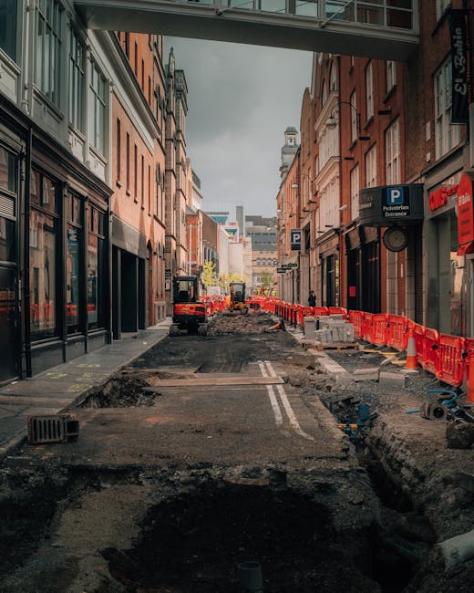 A street in an urban area undergoing roadworks, with an uneven asphalt surface and visible deep trenches for utility installations. Temporary orange safety barriers line the sidewalk on the right side