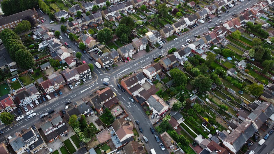 Aerial view of a residential neighbourhood showing a variety of houses with gardens and driveways, arranged along a network of narrow streets. Numerous cars are parked along the roads and within driveways, with some vehicles in motion. The scene includes trees and greenery interspersed among the houses, some backyards featuring swimming pools, patios, and sheds. The image captures the area during daylight with natural lighting. This setting represents a typical suburban street layout suitable for home relocations and furniture transport, as seen in the context of house removals in Ponders End. The presence of vehicles being loaded or unloaded is not visible, but the arrangement suggests a safe environment for moving services like those offered by Man with Van Ponders End, supporting packing, moving, and transport processes involved in house relocations in the EN3 postcode area.