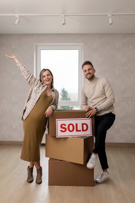 A happy couple stands inside an empty room with a large window, holding a cardboard box labeled 'SOLD' during a home relocation process. The woman, wearing a checked shirt over a beige dress and ankle boots, gestures cheerfully with her right arm extended outward. The man, dressed in a light-colored sweater, dark trousers, and white sneakers, leans on the top box with a smile. The three stacked cardboard boxes are positioned on the wooden floor, indicating they are in the process of packing and preparing for moving services. The room has neutral-toned wallpaper, a white ceiling with track lighting, and natural daylight coming through the window, consistent with an internal setting pre-move, reflecting the final stages of property sale and furniture transport in the context of house removals, as provided by Man with Van Ponders End.