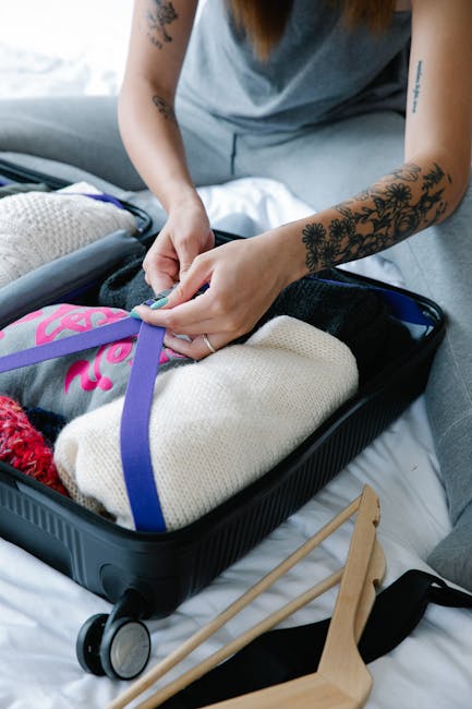 A person with tattooed arms is packing a suitcase on a bed, securing clothing and personal items with a purple strap. The open suitcase contains folded clothing, a cream-colored knitted piece, and other belongings. Nearby on the bed are a pair of wooden slippers with black straps and a trolley handle. The setting appears to be inside a home, with natural light illuminating the scene, illustrating the packing process involved in house removals or home relocation. Occasionally, a moving company like Man with Van Ponders End would assist in such packing and furniture transport activities, especially during the packing tips for flats on Ponders End High Street.