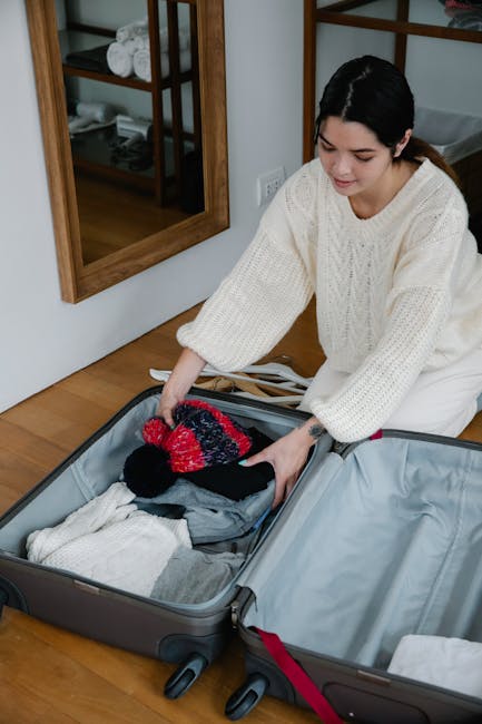 A woman with dark hair, wearing a cream knitted sweater, is seen packing a suitcase on a wooden floor inside a home. She is arranging clothing and accessories, including a colorful knitted hat, into an open, empty suitcase. The room features a large wall mirror with a wooden frame and a shelving unit with rolled towels and folded linens in the background. The suitcase is part of a home relocation or packing process, with additional luggage situated nearby on the floor. This scene depicts the careful organization and preparation involved in house removals or moving services, highlighting packing activities customary in the final stages of a residential move, as handled by companies like Man with Van Ponders End.