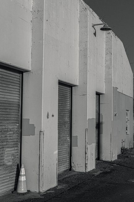 A section of a building exterior showing three metal roller shutters on separate doorways set into a plain concrete wall. The shutters are closed and have visible horizontal slats, with some signs of wear and weathering. One of the doorways has a small orange and white traffic cone placed directly in front of it on the pavement, which appears clean with some minor cracks. An outdoor shower head fixture is mounted high on the wall above the middle doorway, and a small bush is visible near the base of the wall on the right side. The lighting suggests daytime with shadows cast along the wall, and the environment looks like a loading or storage area. Occasionally, a move company like Man with Van Ponders End might use such exterior spaces for loading or moving furniture during a home relocation or packing and moving process, especially near loading bays and permits areas explained for efficient vehicle access.