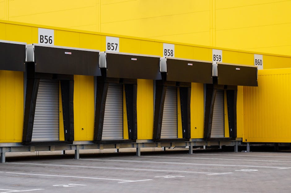 The image shows a section of a yellow loading bay structure with five designated bays labeled B56, B57, B58, B59, and B60 in black text on white signage. Each bay has a black retractable shutter door with beige corrugated surfaces, situated inside an industrial or commercial building. The bays are supported by black metal brackets and are set above a pavement area with white parking lines, indicating an outdoor or semi-outdoor loading zone. These bays are designed for vehicle access to facilitate home relocation services, furniture transport, and packing and moving tasks. The lighting appears natural, casting minimal shadows, with the yellow colour of the structure contrasting with the darker elements of the doors. The setting highlights a professional vehicle loading area managed by [COMPANY_NAME], suitable for efficient removal and logistics operations related to house removals and moving logistics.