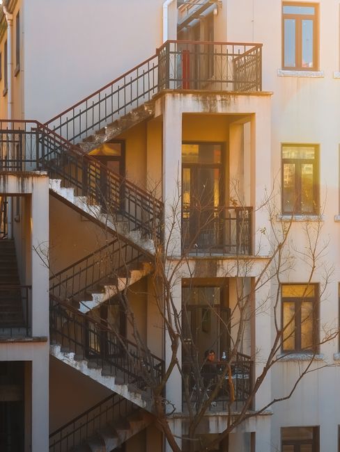 The exterior of a multi-storey residential building with an external staircase made of concrete and metal railings, leading to balconies on each floor. The building's facade features several windows and doors, with some balconies decorated with small plants and furniture. The staircase appears slightly weathered, with some paint chipping visible. A leafless tree is situated close to the building, partially obscuring the lower part of the structure. The scene is lit by natural daylight, suggesting daytime conditions. This image captures a typical urban residential environment, relevant to house removals and moving services, illustrating the logistical considerations of navigating staircases during a home relocation managed by Man with Van Ponders End.