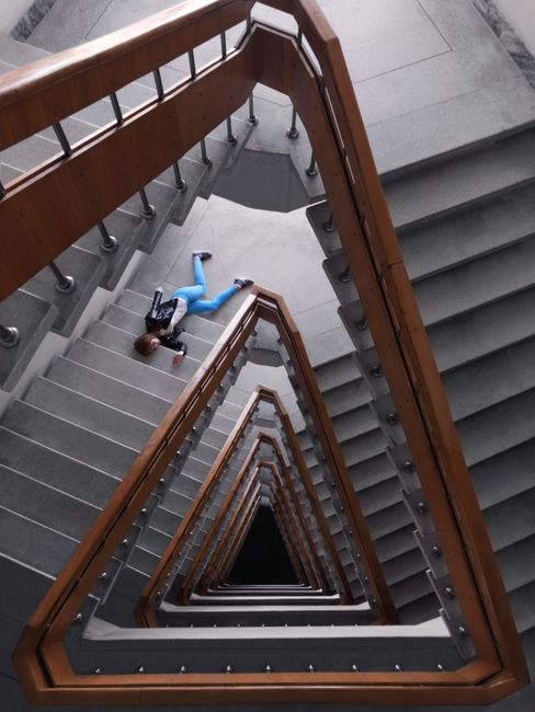 A view looking down a spiral staircase inside a building, showing multiple landings with grey steps, wooden handrails, and metal balusters. A woman dressed in a blue sweater and dark pants is lying on one of the landings, appearing to rest or pose. The staircase creates a geometric, tunnel-like perspective extending towards a black central opening at the bottom. The surrounding environment includes grey tiled floors on each landing, and the lighting appears natural, illuminating the staircase evenly. This image exemplifies a typical interior space involved in household relocation or moving logistics, where staircases may present challenges during furniture transport. Man with Van Ponders End regularly assists with such home removals, including navigating confined staircases during the loading and unloading process.